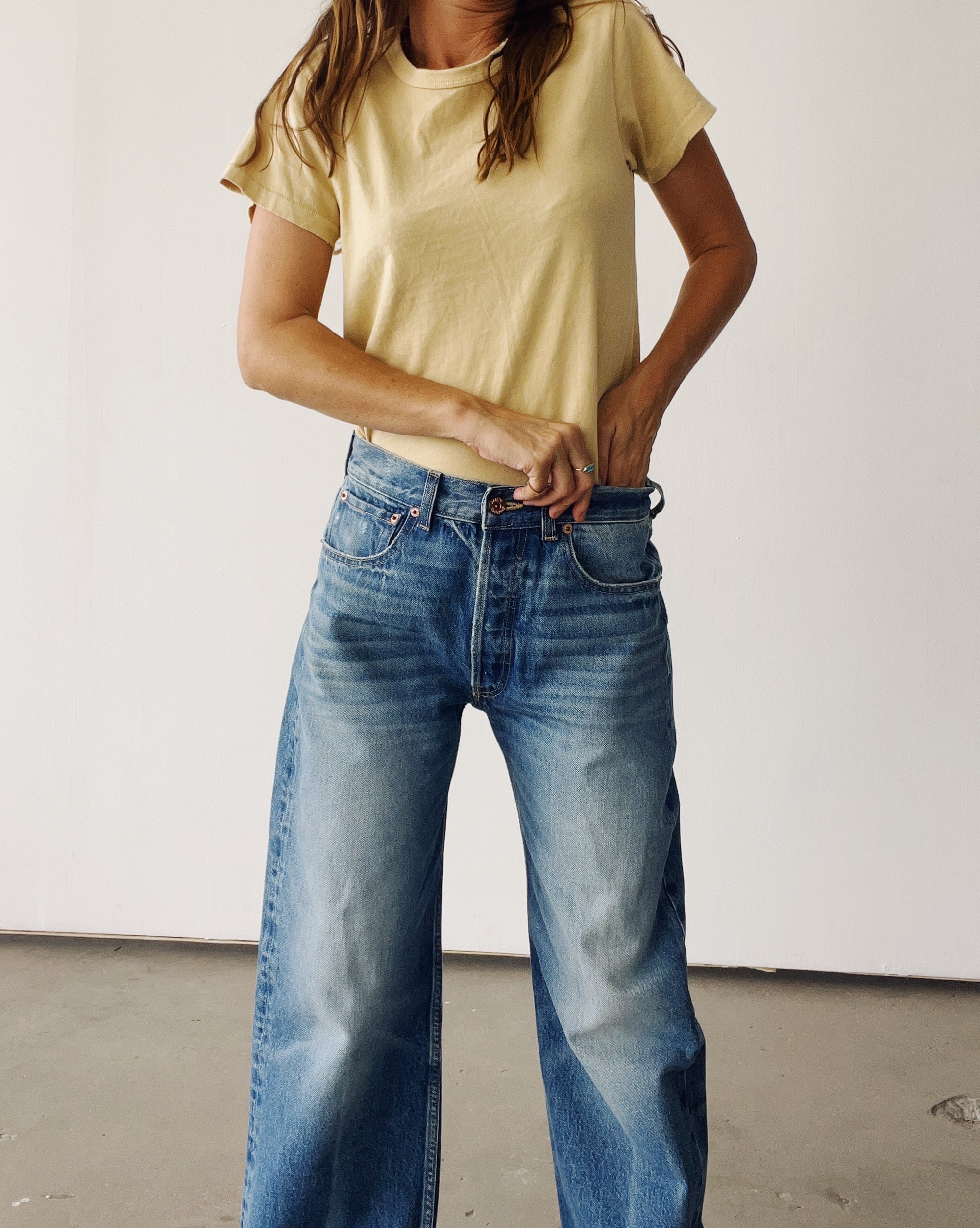 A person indoors fastens the waistband of blue vintage-fit jeans while wearing Hey Gang's The Crew 26, a yellow 100% cotton t-shirt. Their face is out of frame against a plain light-colored background.