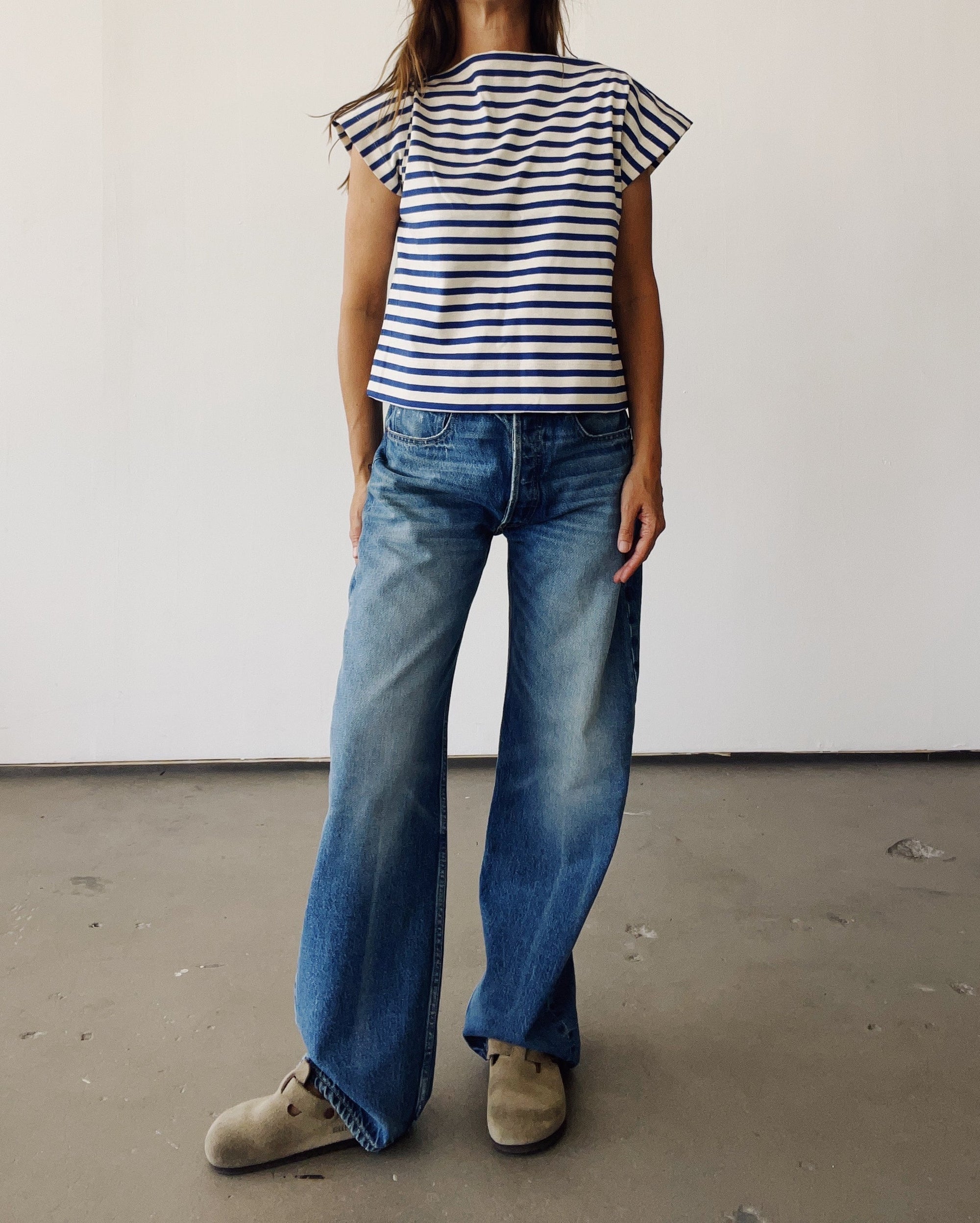 A person stands indoors wearing Hey Gang's The Sailor Shirt in black and white Japanese cotton, paired with wide-leg blue jeans and beige shoes. The plain background and cropped frame hide the person's head.