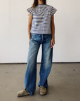 A person stands indoors wearing Hey Gang's The Sailor Shirt in black and white Japanese cotton, paired with wide-leg blue jeans and beige shoes. The plain background and cropped frame hide the person's head.