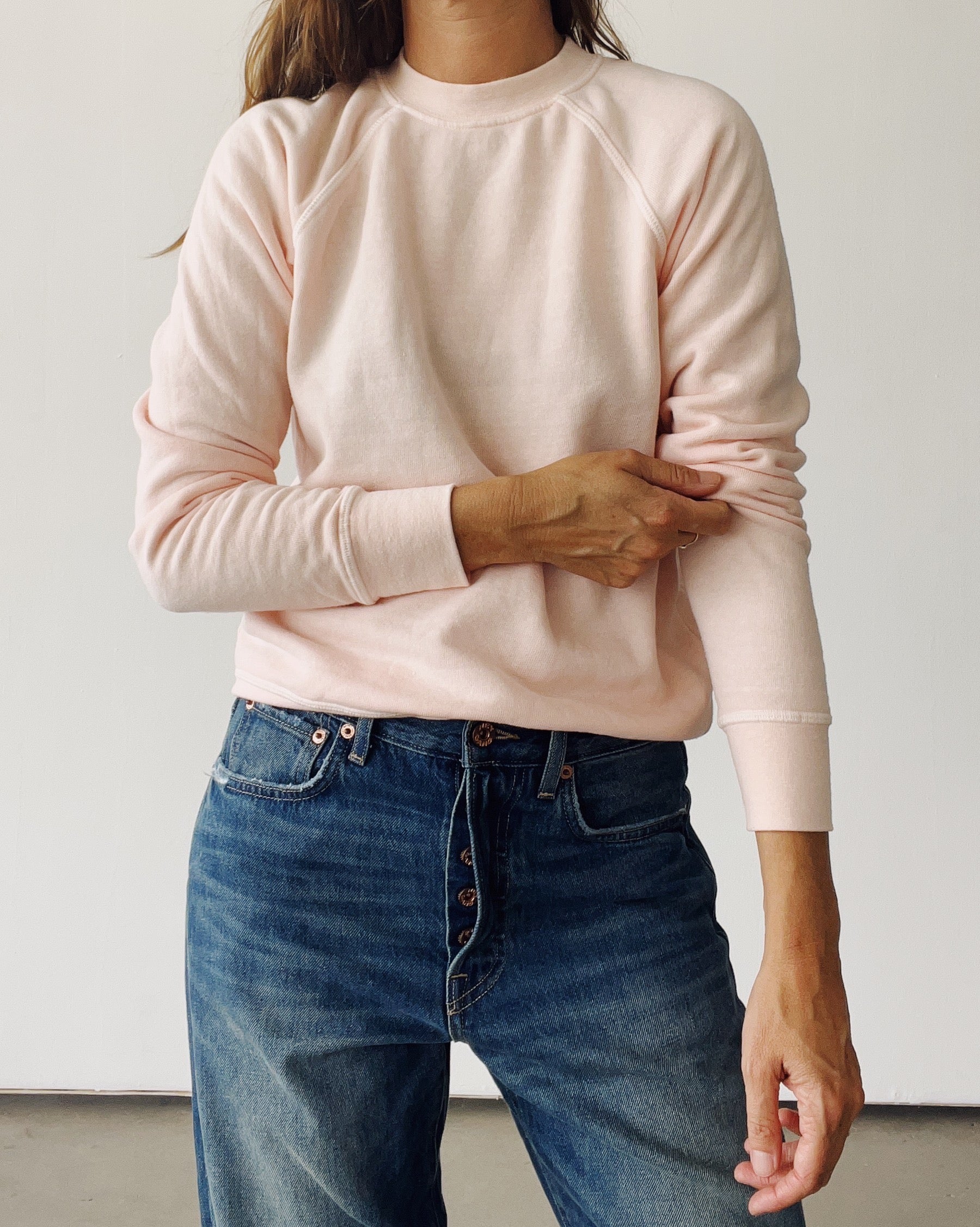 A person wearing Hey Gang’s The Sweatshirt 26 in light pink tri-blend fleece and blue jeans stands against a white background, adjusting their sleeve. Their face is not shown.