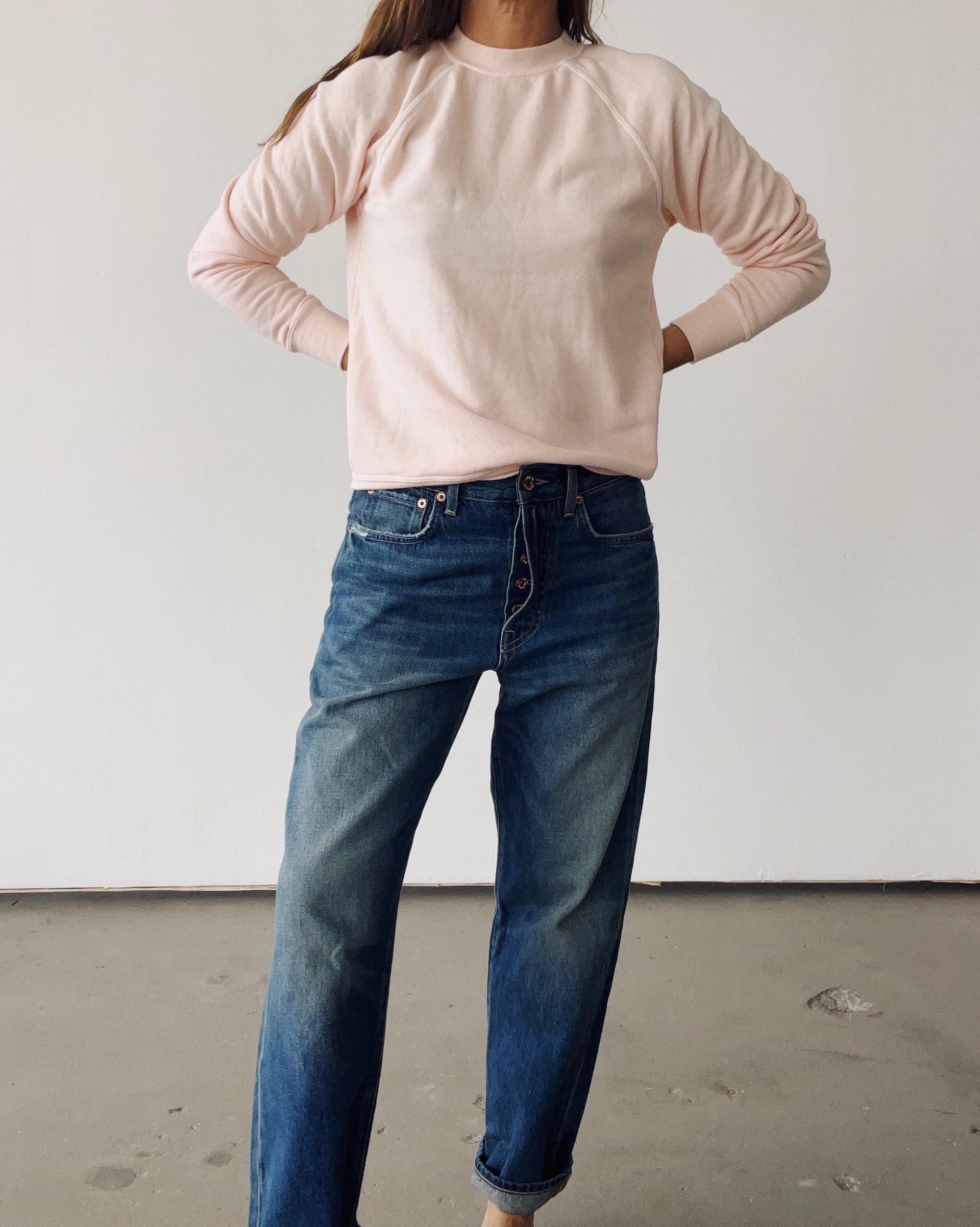 A person stands indoors against a plain white wall, wearing Hey Gang's The Sweatshirt 26 in light pink and blue jeans, hands on hips. Their head is not visible in the image.