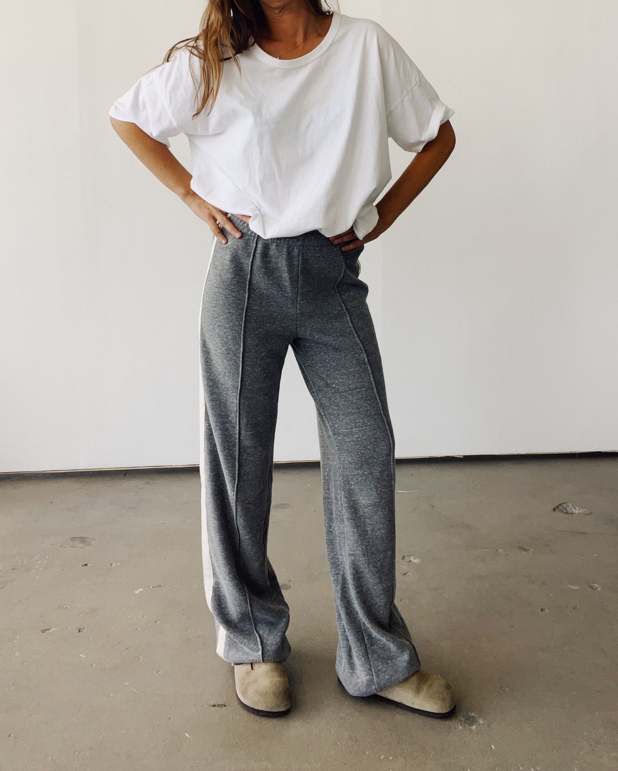 Indoors, a person wears Hey Gang's The Track Pant—high-waisted, gray, wide leg, tri-blend fleece—paired with a loose white tee and tan slippers. Their hands are on their hips; their head is not visible.