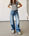 Indoors against a minimal backdrop, a person wears Hey Gang’s The Wide Slouch Jean in Harris wash, styled with a grey t-shirt and beige slip-ons. Hands on hips, their face is out of frame.