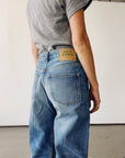 Someone wearing The Wide Slouch Jean by Hey Gang stands with their back to the camera, showing the tan "HEY GANG" label above the back pocket. They also wear a gray t-shirt. The background is plain and light-colored.