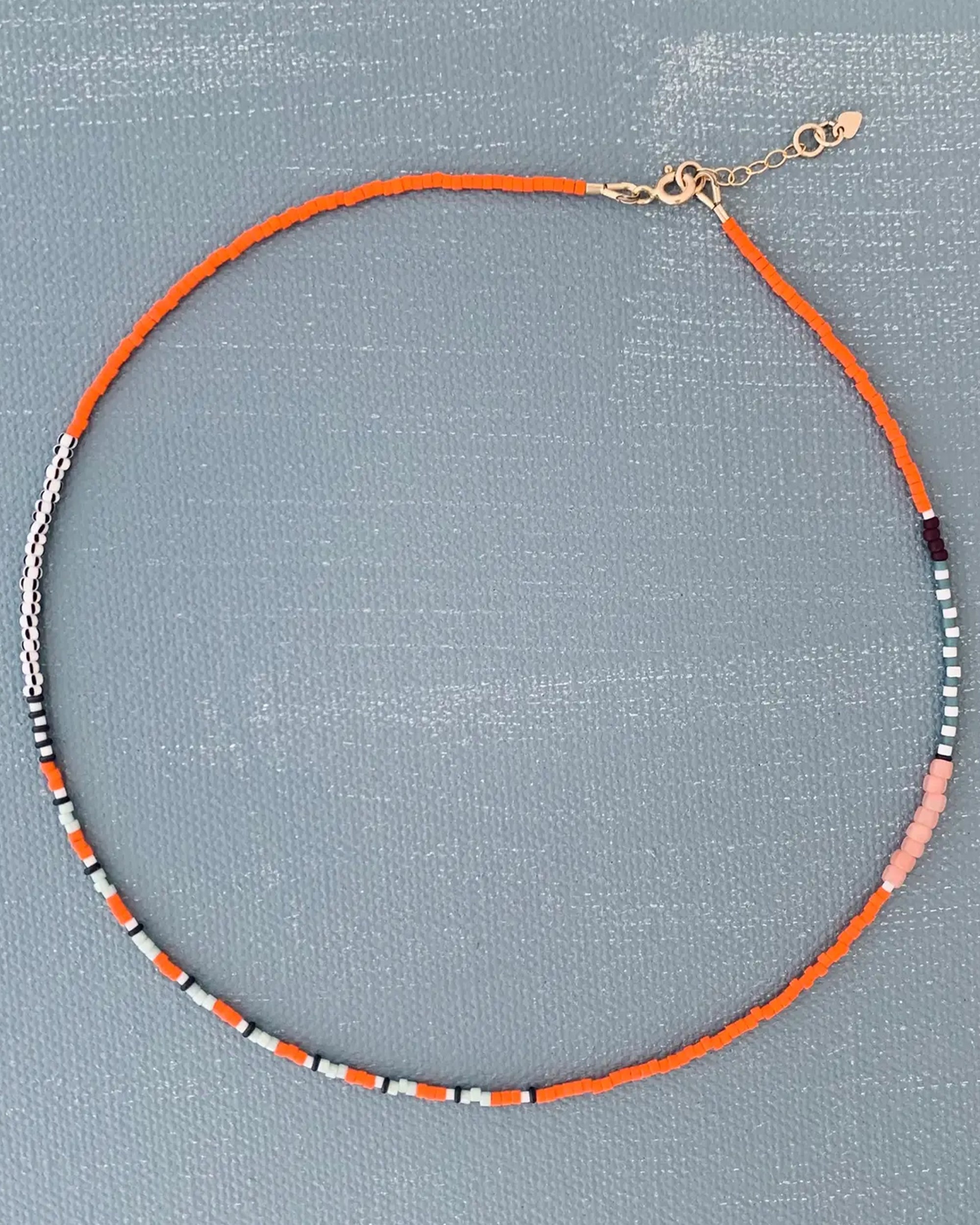 The AR Line Necklace in Marigold by Faire features Japanese glass beads in orange, white, black, and pink with a gold-filled clasp and adjustable chain, shown on a textured gray surface.