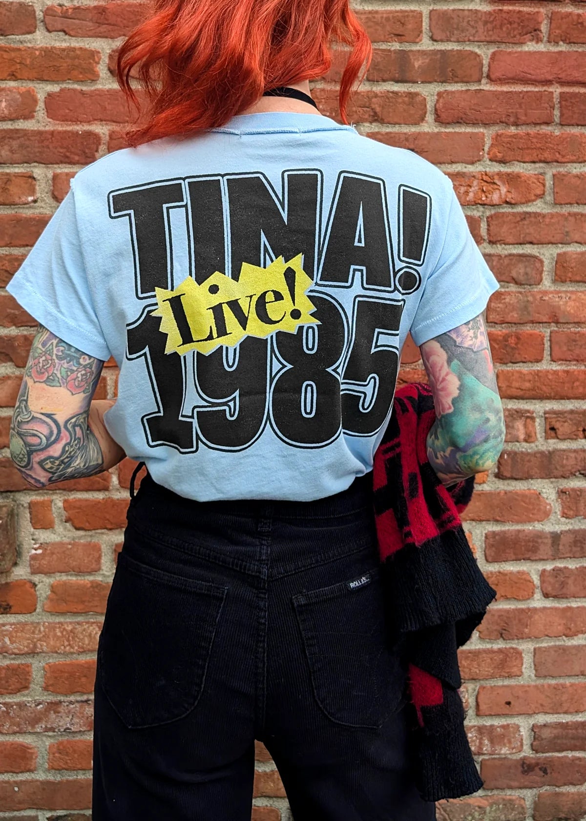 A person with red hair and colorful tattoos faces a brick wall, wearing the DayDreamer Tina Turner 1985 Reverse DF Tee, which features bold "TINA! 1985 Live!" text on the back. They’re holding a black and red scarf.