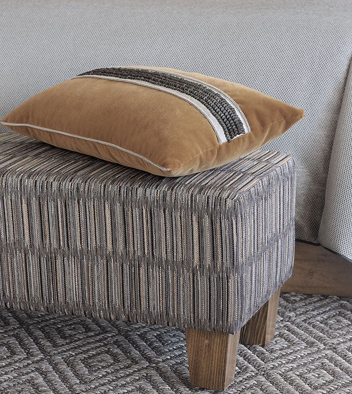 The Eastern Accents TAOS FAUX MOHAIR pillow, featuring two dark textured stripes and subtle beaded trim, sits on a patterned upholstered ottoman with wooden legs beside a gray textured surface and woven rug.