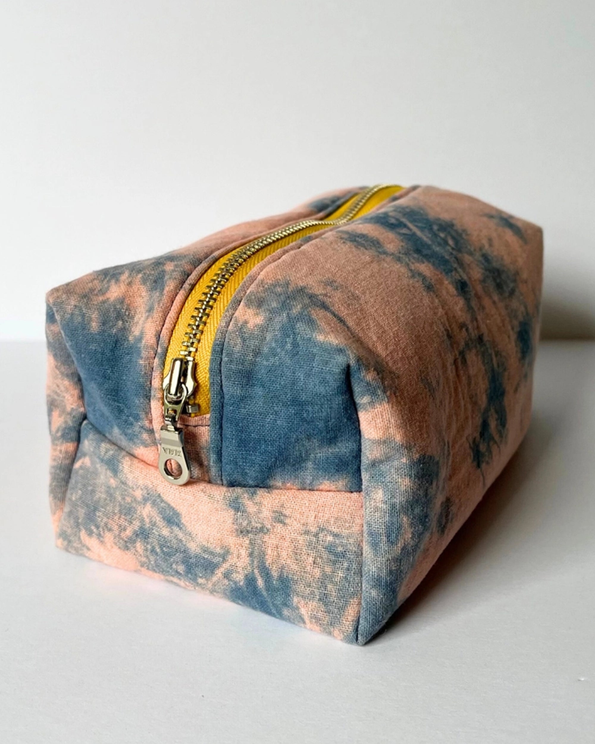 The Faire Dyed Toiletries Bag, made from 100% cotton with a yellow zipper and hand-dyed blue and peach tie-dye design, is displayed on a plain white surface.