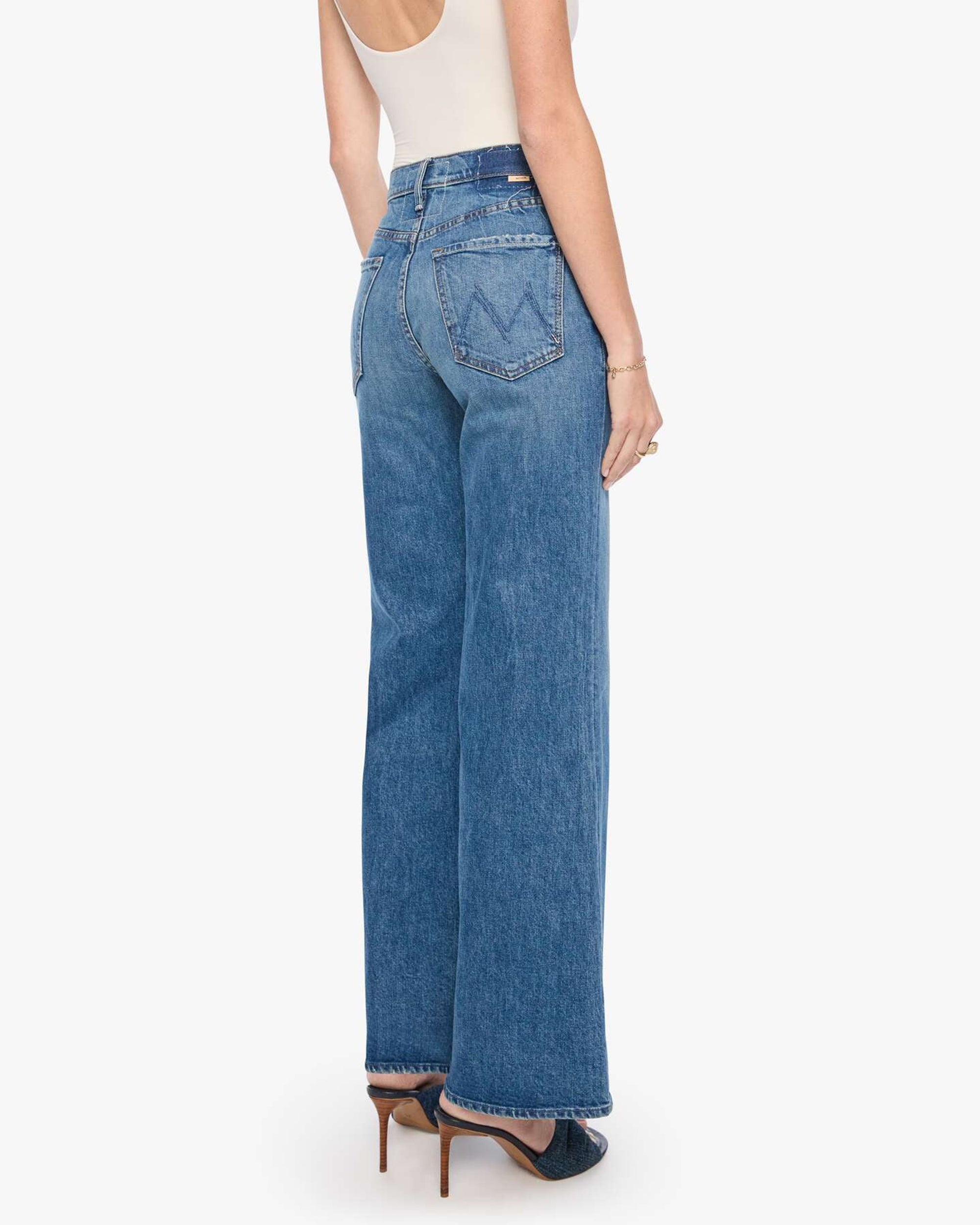 The Tomcat Roller Sneak jeans by Mother in blue denim, styled with a white tank and heels, shown from the back.