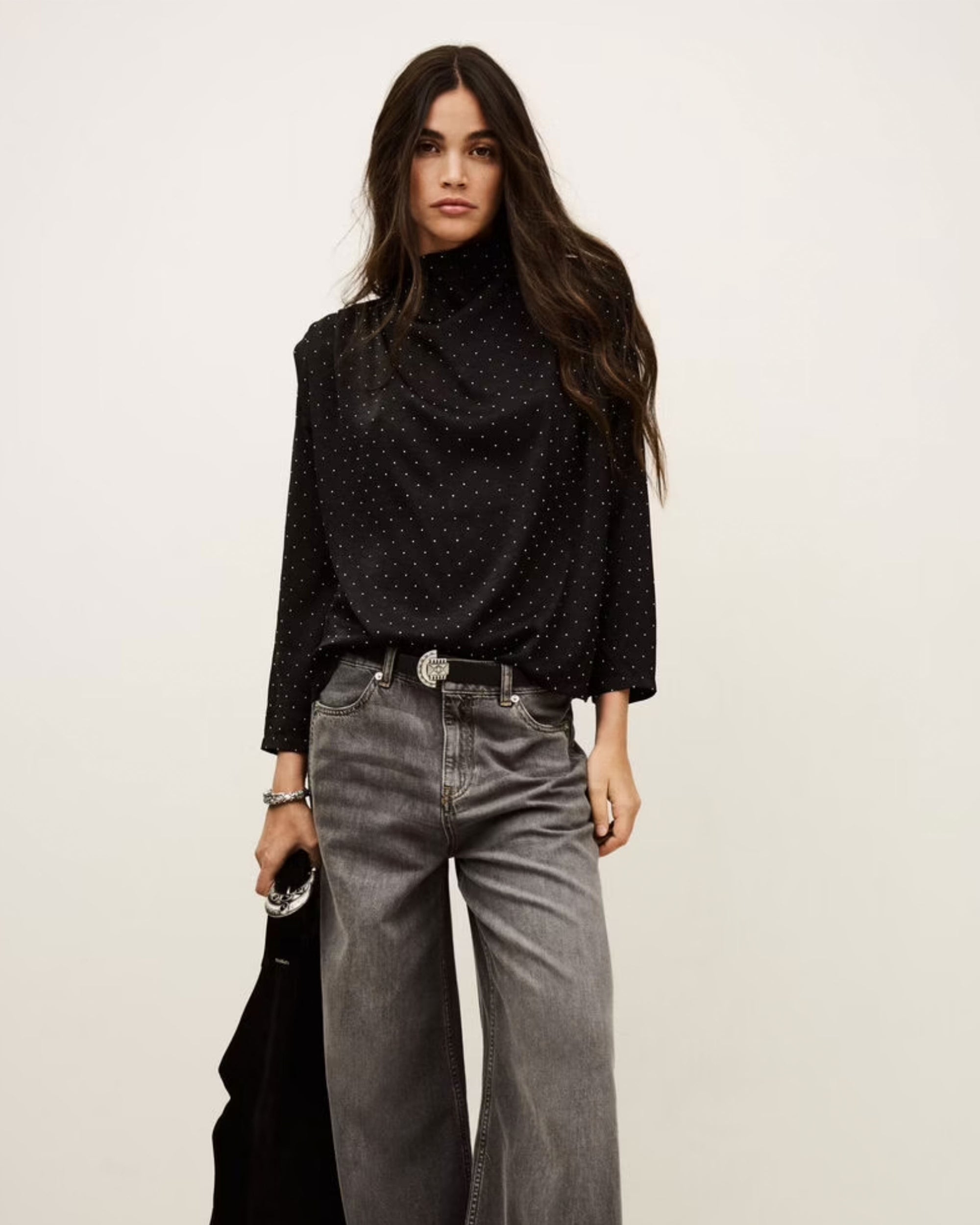 A woman with long dark hair wears the BA&SH Top Debra, a black high-neck polka-dot blouse with batwing sleeves, and loose gray jeans. She holds a black jacket and stands against a plain light background.