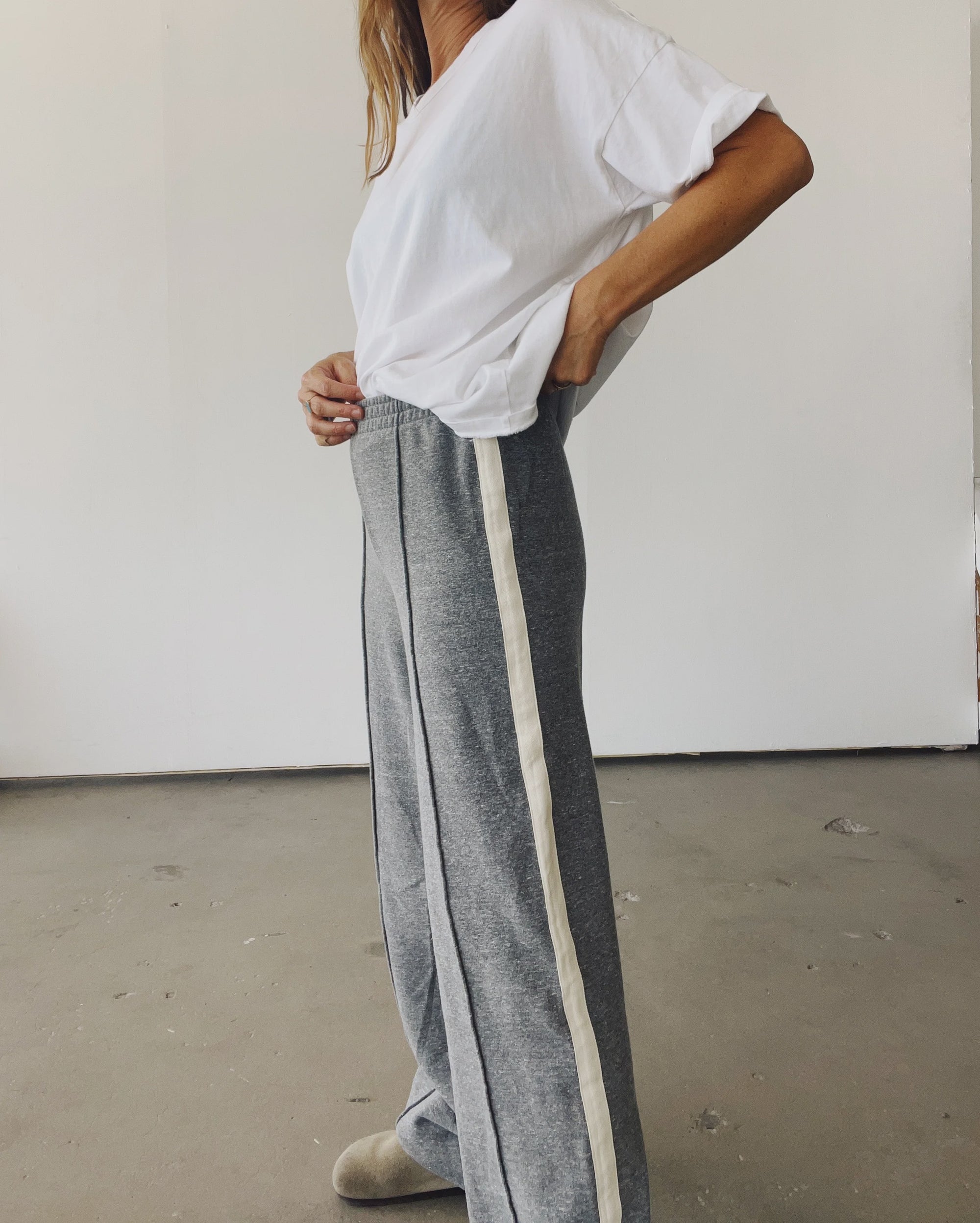 A person stands indoors, hands on hips, wearing Hey Gang’s The Track Pant—gray wide-leg pants with a white side stripe—and a loose white t-shirt against a plain, minimalistic background.