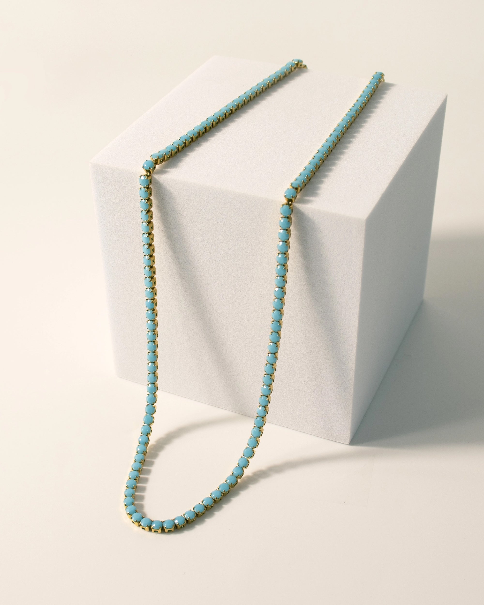 The Faire Turquoise Tennis Necklace features turquoise rhinestones on a hypoallergenic, 18K Gold PVD coated stainless steel chain, draped over a white cube and displayed against a light, neutral background.