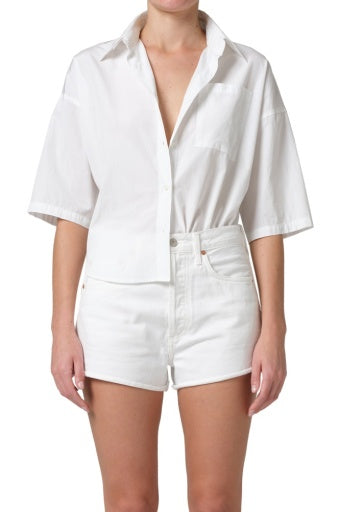 A model wears the Frances Cropped Cape Button Up by Citizens Of Humanity/AGOLDE—100% cotton, loose fit, short sleeves—paired with high-waisted white shorts, standing against a plain white background for a modern take on classic style.