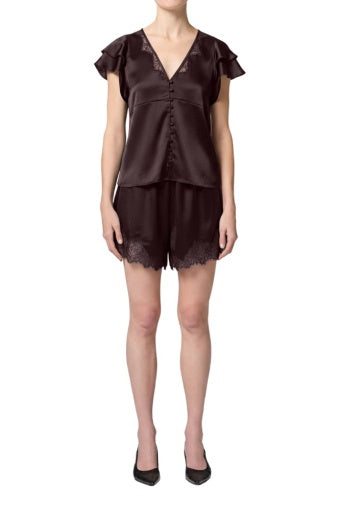 A woman wears Citizens Of Humanity/AGOLDE's Lace Trimmed Tap Short—a dark brown satin pajama set with lace trim, short-sleeve V-neck buttoned top, and matching shorts—paired with black heels against a white background.