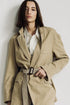 A woman with straight brown hair wears the Denimist DECONSTRUCTED BLAZER WITH BELT in beige over a white pinstriped shirt, standing against a plain light background and looking at the camera with a neutral expression.