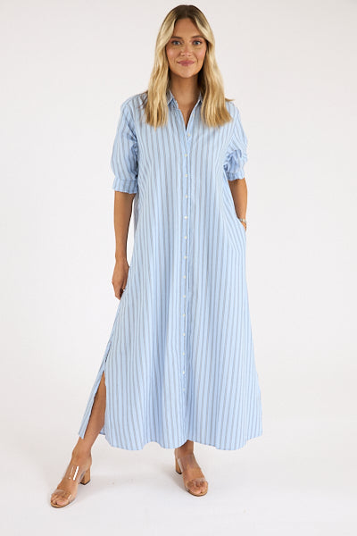 A woman poses against a plain background wearing the Xirena Boden Dress, a light blue, ankle-length cotton poplin shirtdress with elbow-length sleeves and side slits, styled with nude heeled sandals.