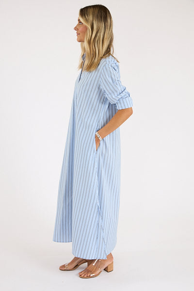 A woman in profile wears the Xirena Boden Dress—a long, light blue cotton poplin shirtdress with vertical white stripes. She has wavy, shoulder-length blonde hair, nude heeled sandals, and her hands in the dress pockets.