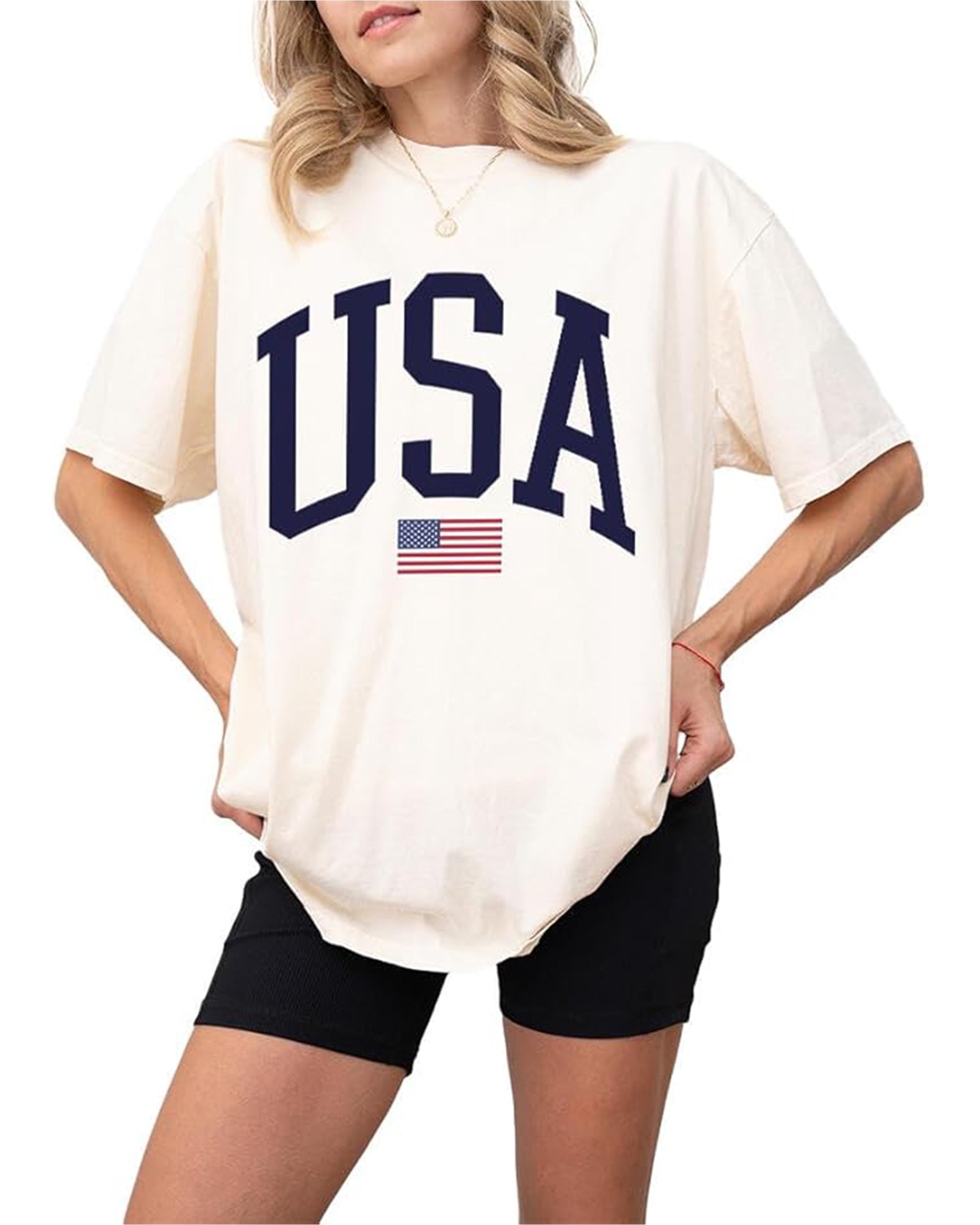 A person wears the Faire USA Flag Tee, a loose cotton shirt with "USA" in bold black letters and a small flag beneath, paired with black shorts and hands in pockets for an effortlessly comfortable look.