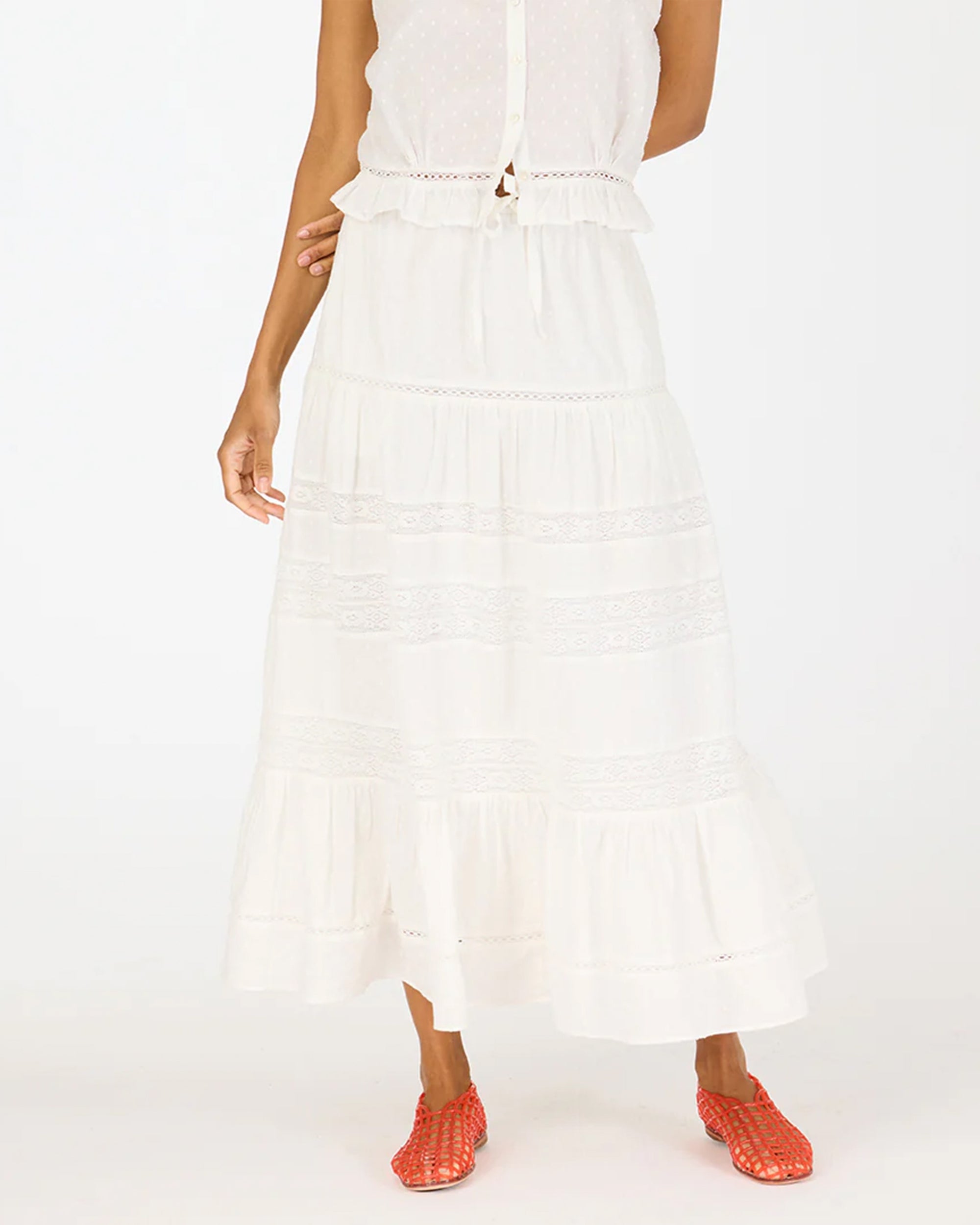 Wearing a sleeveless white top and the Kerri Rosenthal Vacance Skirt with tiered lace trim, this person stands against a plain background, finishing the outfit with bold red woven shoes.