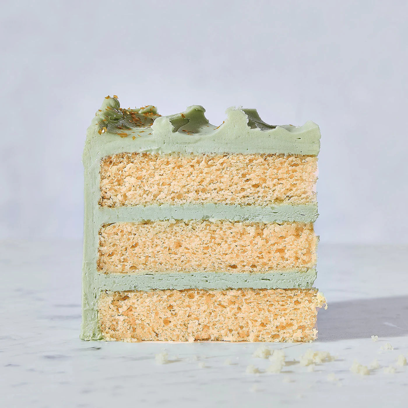 A slice of three-layer vanilla cake made with Wild Rye Cake Mix, featuring light green frosting between layers and on top, sits on a light surface with a few cake crumbs nearby—perfect for vanilla lovers.