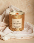 A scented candle labeled "Velvet Cowboy" by Faire, with a single wick, displayed on a soft, crumpled fabric against a marbled beige background. The FEBE candle's container has a speckled gold Arizona-style design.