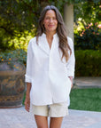 A woman with long brown hair smiles warmly outdoors, hands in her pockets, wearing the Frank & Eileen Victoria Collarless shirt and light-colored shorts, standing amid garden greenery and a large decorative vase.