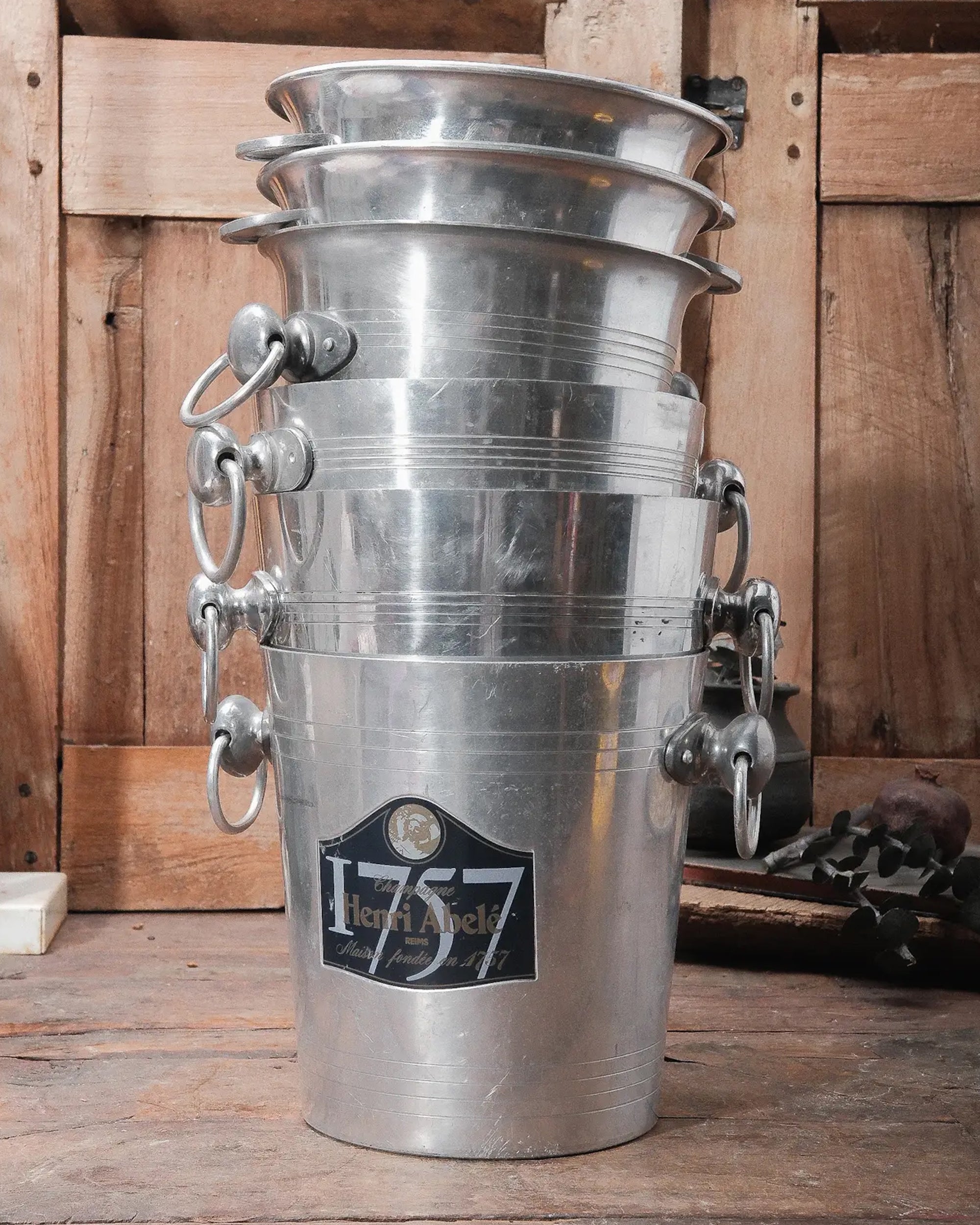 A stack of five Go Home Vintage Champagne Buckets with handles sits on a rustic wooden table; the top bucket features a "75 Henri Abelé" label.