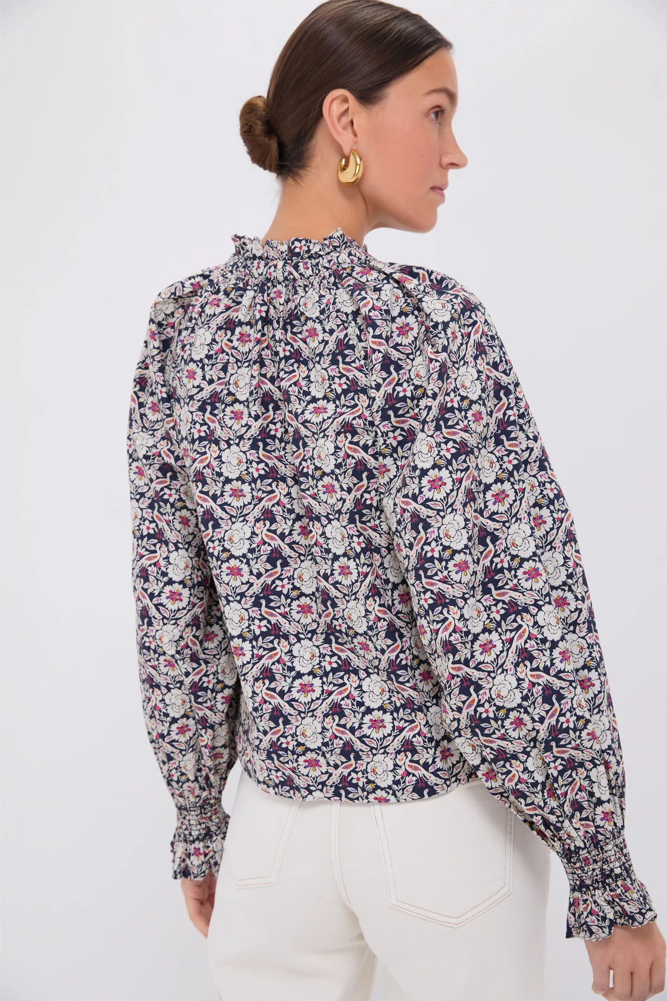 A person with dark hair in a bun models Rhode's Aiden Top, adorned with floral patterns, a ruffle collar, and self-tie bows on long sleeves. They pair it with white pants and stand against a plain white background, looking to the side.