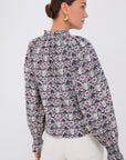 A person with dark hair in a bun models Rhode's Aiden Top, adorned with floral patterns, a ruffle collar, and self-tie bows on long sleeves. They pair it with white pants and stand against a plain white background, looking to the side.