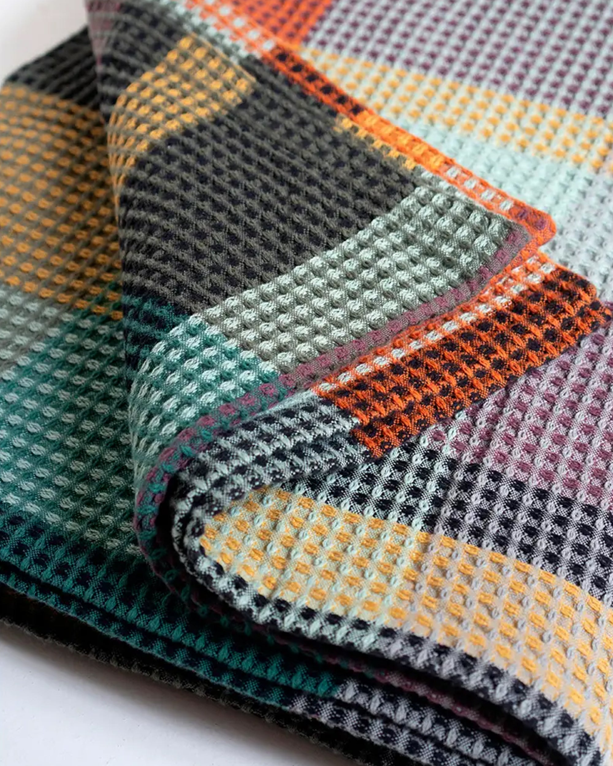 A close-up of the Faire Vrou-Vrou Blanket, folded to show its waffle texture and vibrant geometric pattern in orange, yellow, green, black, and purple for a modern look.