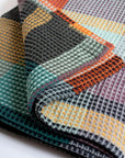 A close-up of the Faire Vrou-Vrou Blanket, folded to show its waffle texture and vibrant geometric pattern in orange, yellow, green, black, and purple for a modern look.