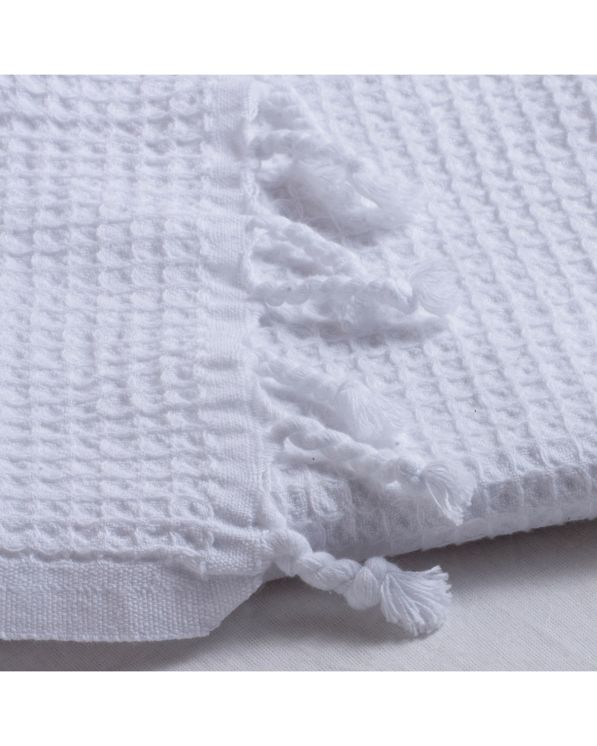 A close-up of the Faire Waffle Hand Towel, made from white Turkish cotton with a waffle weave and decorative twisted fringe tassels, displayed on a white surface.