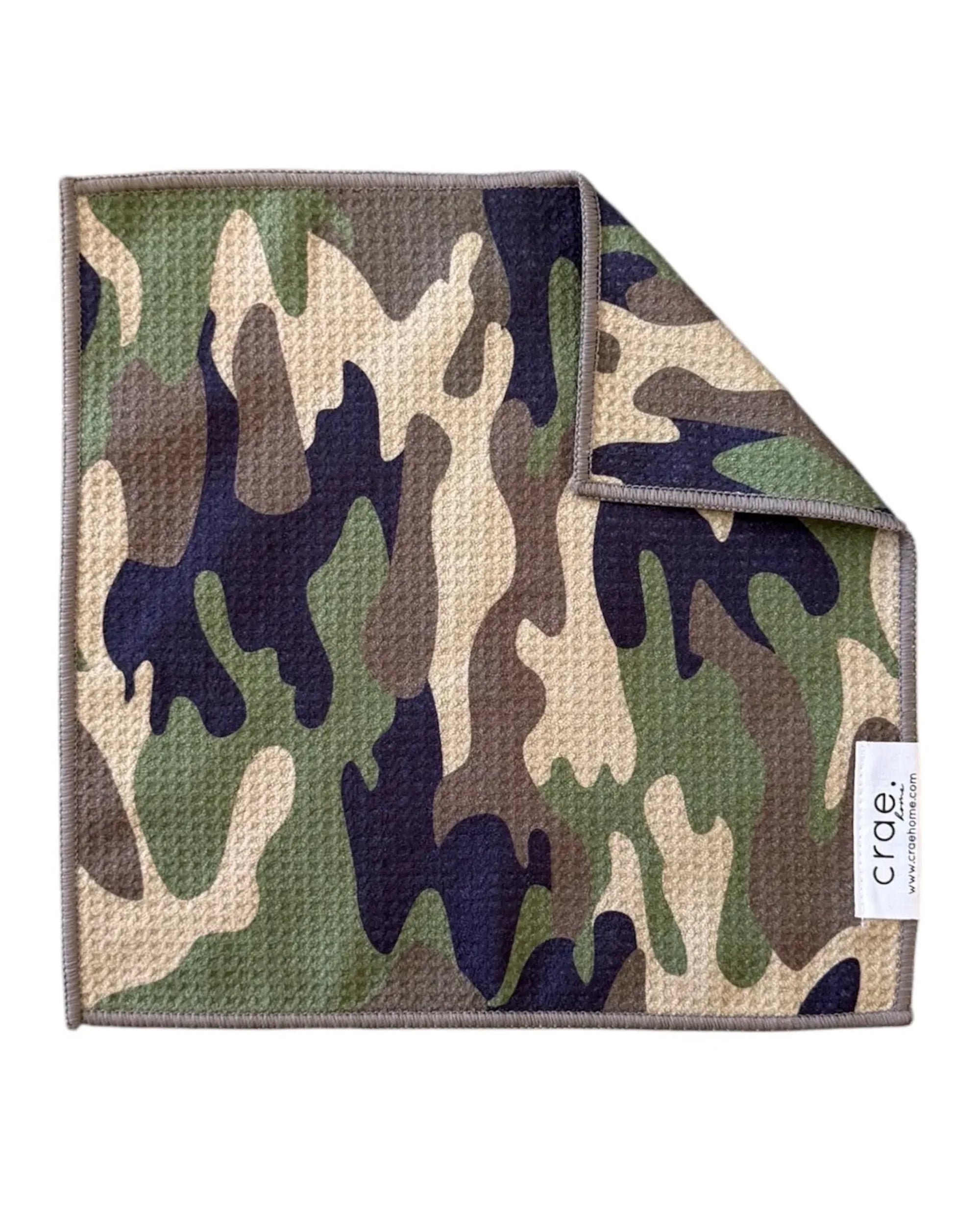 A square waffle weave microfiber washcloth from Faire, featuring a green, brown, tan, and black camouflage pattern with a stitched border. Highly absorbent and eco-friendly. One corner is slightly folded over.