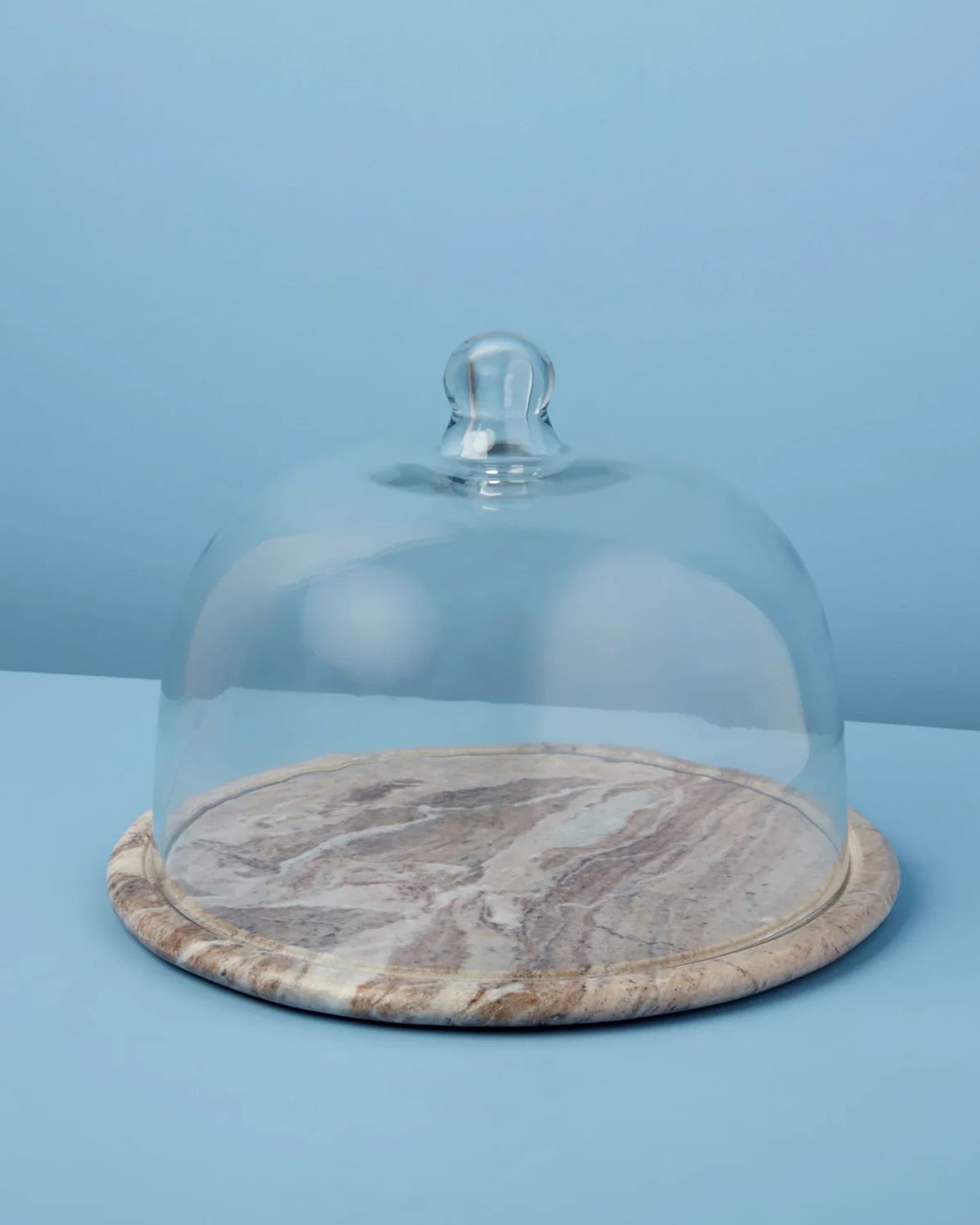 The Be Home 58-377 Waterfall Marble Cloche features a glass dome with a top knob over an empty, round, hand-finished marble base, displayed on a light blue background.