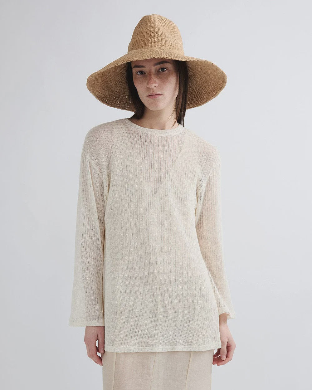 A person in the Janessa Leone Waverly Sand raffia straw hat with a shapeable brim wears a sheer, long-sleeved off-white top, standing against a plain light background and looking at the camera neutrally.