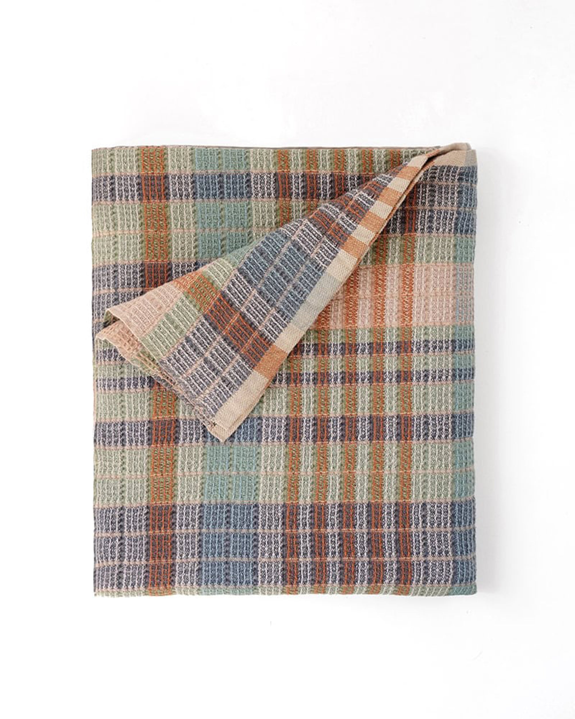 The Faire Wayfarer Towel, featuring a textured plaid pattern in muted green, blue, orange, and beige tones, is shown folded with one corner turned over to display the reverse side against a white background.