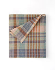 The Faire Wayfarer Towel, featuring a textured plaid pattern in muted green, blue, orange, and beige tones, is shown folded with one corner turned over to display the reverse side against a white background.