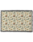 The Faire Western Blanket is a rectangular woven throw with fringe, showcasing cowboys on horseback, green cacti, campfires, and desert plants in a vintage-inspired pattern on a cream background.