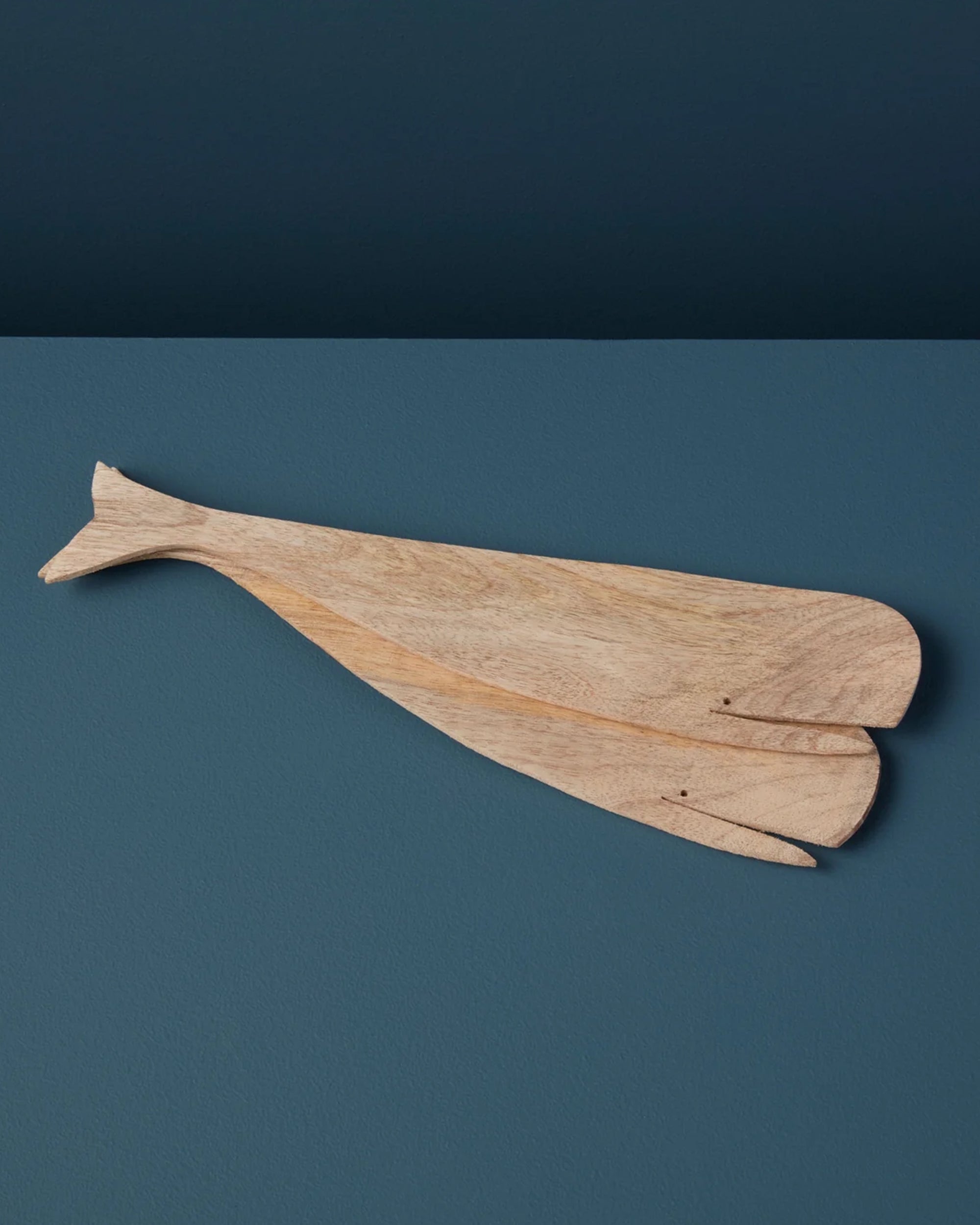 The Sydney Whale Serving Set by Be Home is a handcrafted mango wood cutting board, shaped like a whale with carved tail and mouth details, sustainably sourced and displayed against a dark blue background.