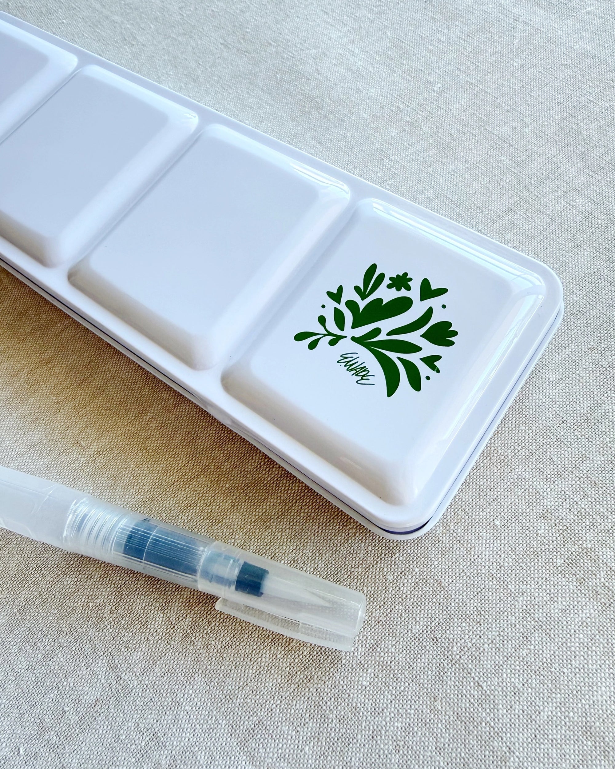 The Faire Watercolor Paint Set w/ Brush, featuring a white metal tin with a green leafy "allegro" design and a clear water brush pen, rests on light beige fabric.