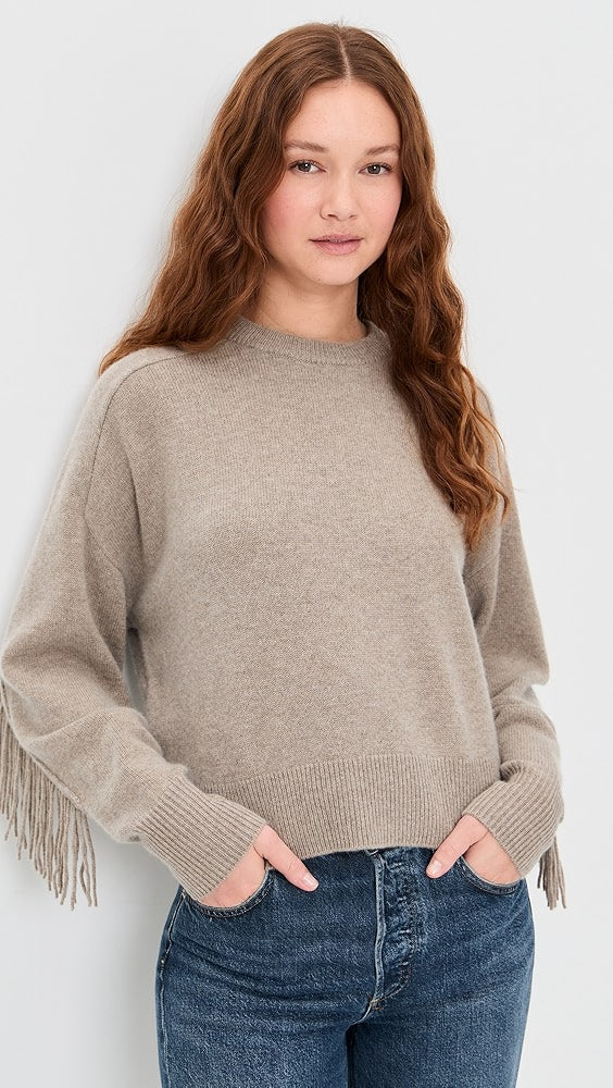 A young woman with long, wavy, auburn hair wears White + Warren’s Cashmere Luxe Fringe Crew in beige and blue jeans, standing against a light background with her hands in her pockets.