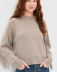 A young woman with long, wavy, auburn hair wears White + Warren’s Cashmere Luxe Fringe Crew in beige and blue jeans, standing against a light background with her hands in her pockets.