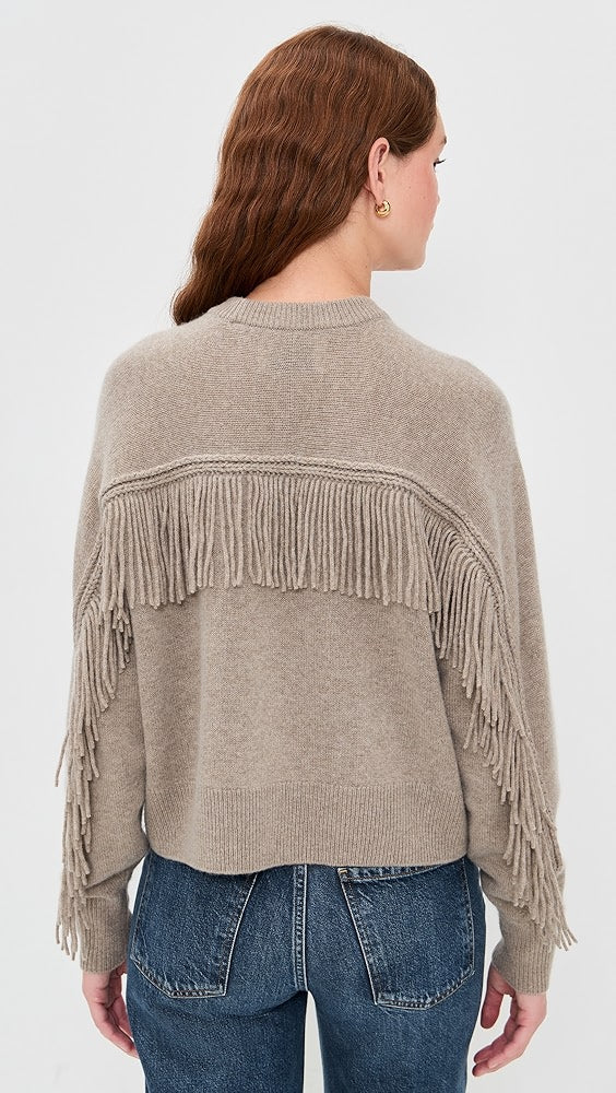 A person with long, wavy brown hair is seen from behind wearing the White + Warren Cashmere Luxe Fringe Crew in taupe, paired with blue jeans. The oversized sweater features fringe on the back and sleeves, set against a light-colored background.
