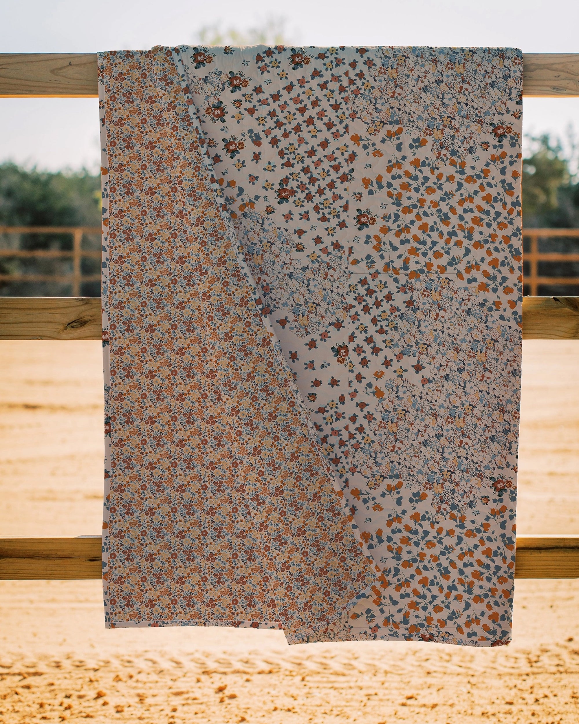 The Winchester Orange Quilt by Faire, ideal for cozy quilts, showcases three sections of colorful floral patterns and is displayed draped over a wooden fence outdoors with sandy ground and blurred trees in the background.