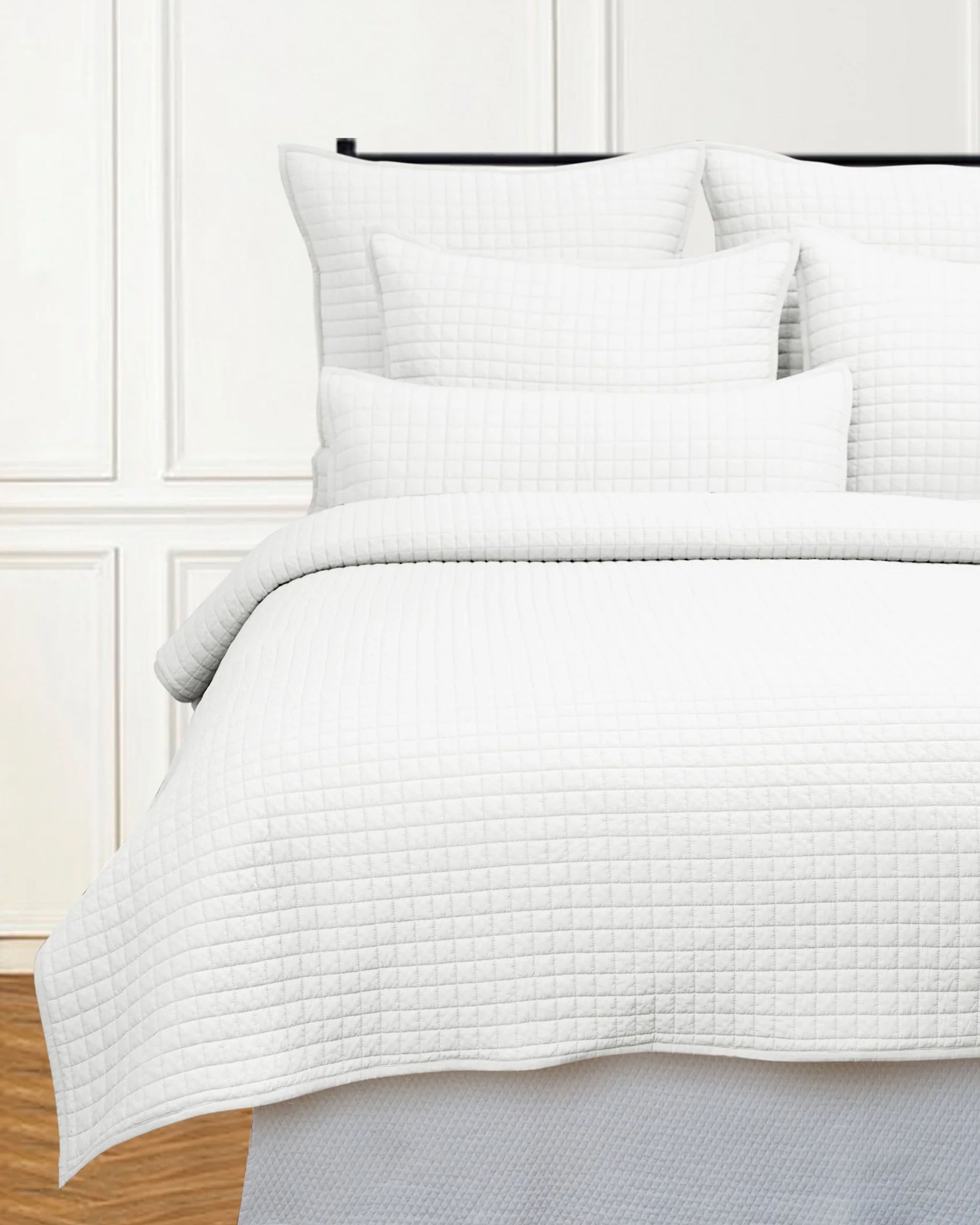 A queen bed is styled with Avasa Bedding's Willa Quilt White Queen and matching pillows, featuring a subtle checkered pattern, all set against a white paneled wall.