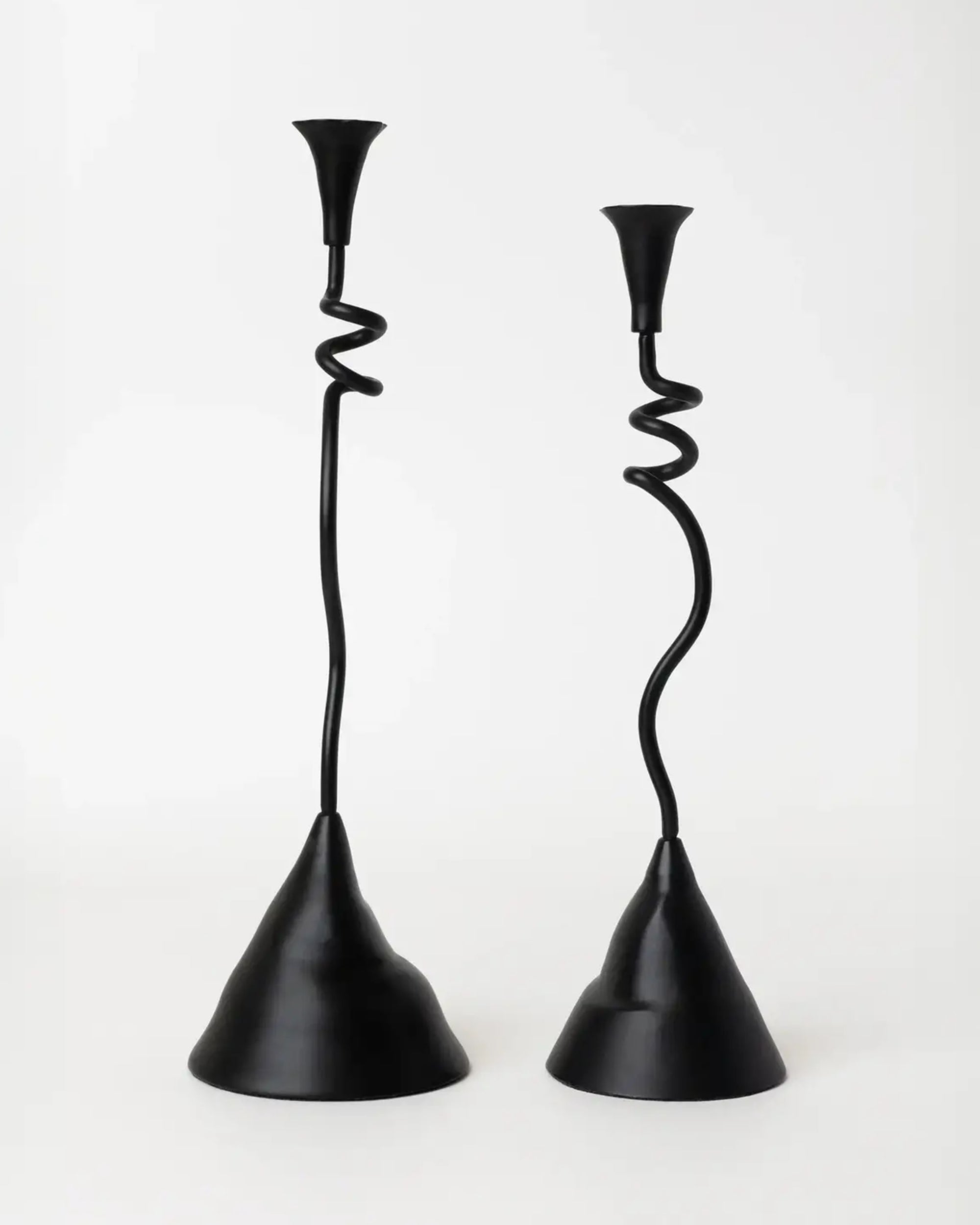 The Faire Winding Taper Candle Holders feature black powder-finished aluminum with twisted, wavy stems and wide, uneven bases. Their unique, sculptural design adds a modern touch to any décor. Set of two shown against a plain white background.