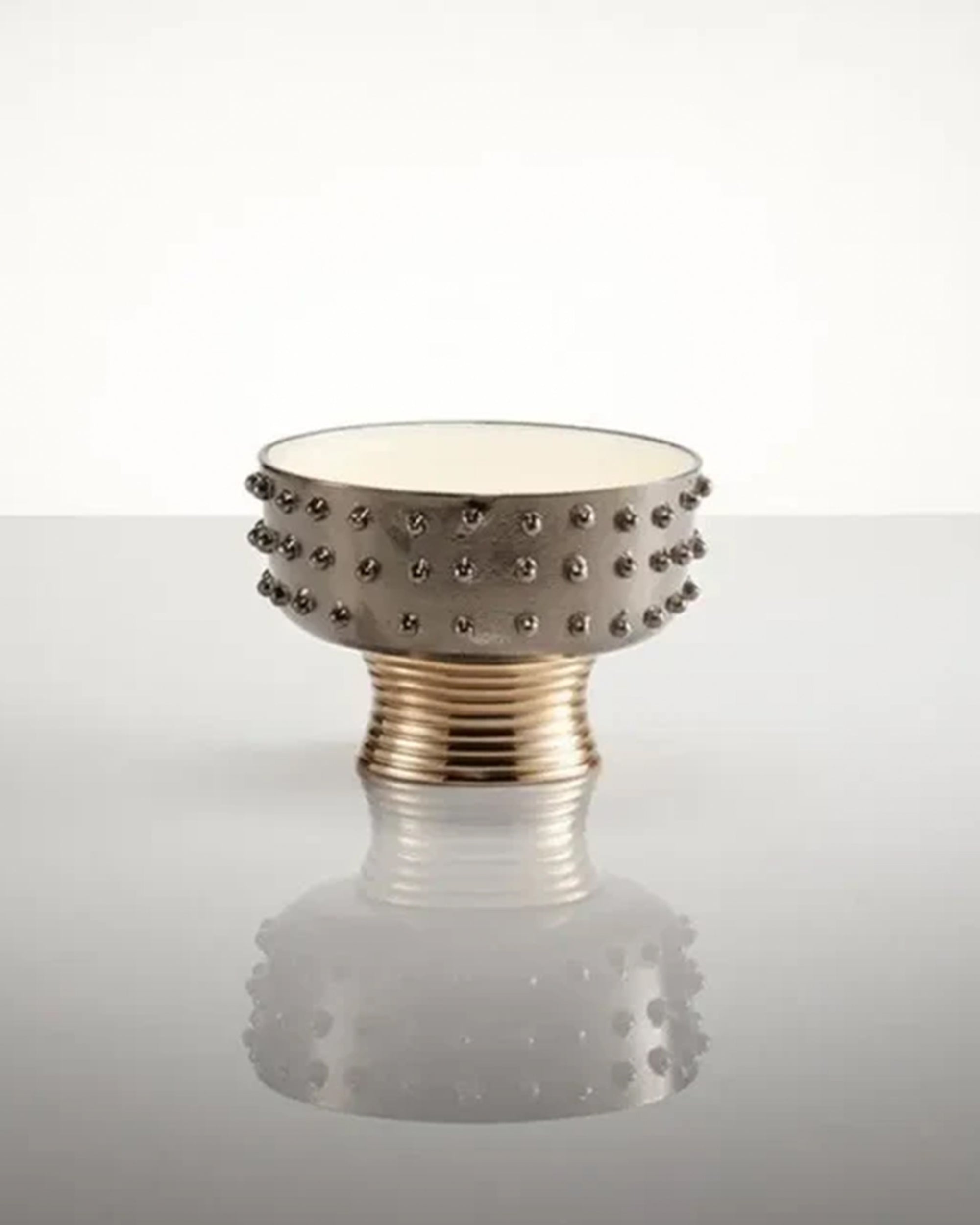 The Faire Djembe Wine Caddy/Nut Dish is a metallic, round bowl with a wide opening, small studs around the middle, and a ribbed base. It rests on a reflective surface against a plain white background.