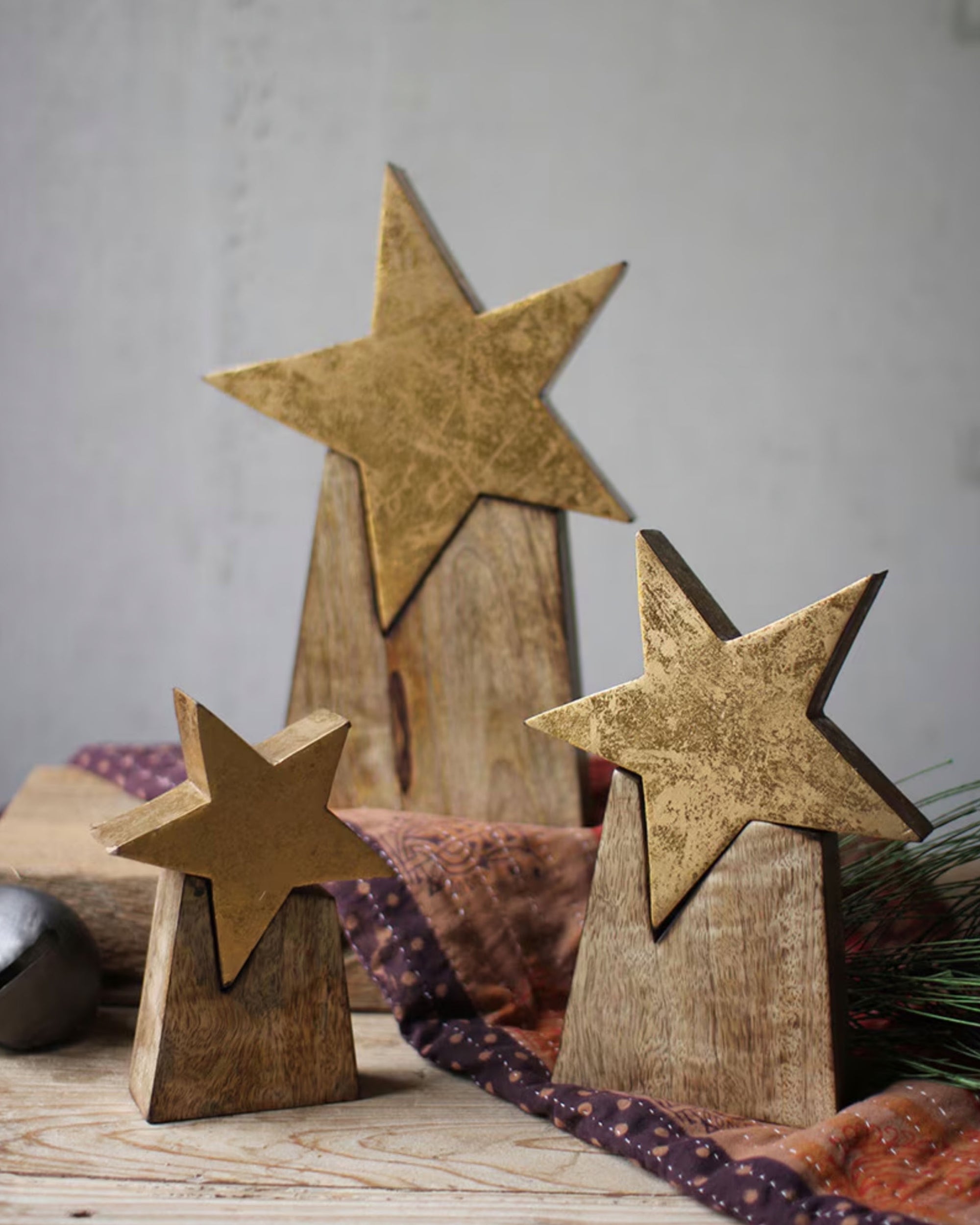 Three Kalalou, Inc NNV1008K Wooden Star On Base decor pieces in gold finish stand at different heights on a patterned cloth, paired with a silver ball and greenery for a festive holiday display.