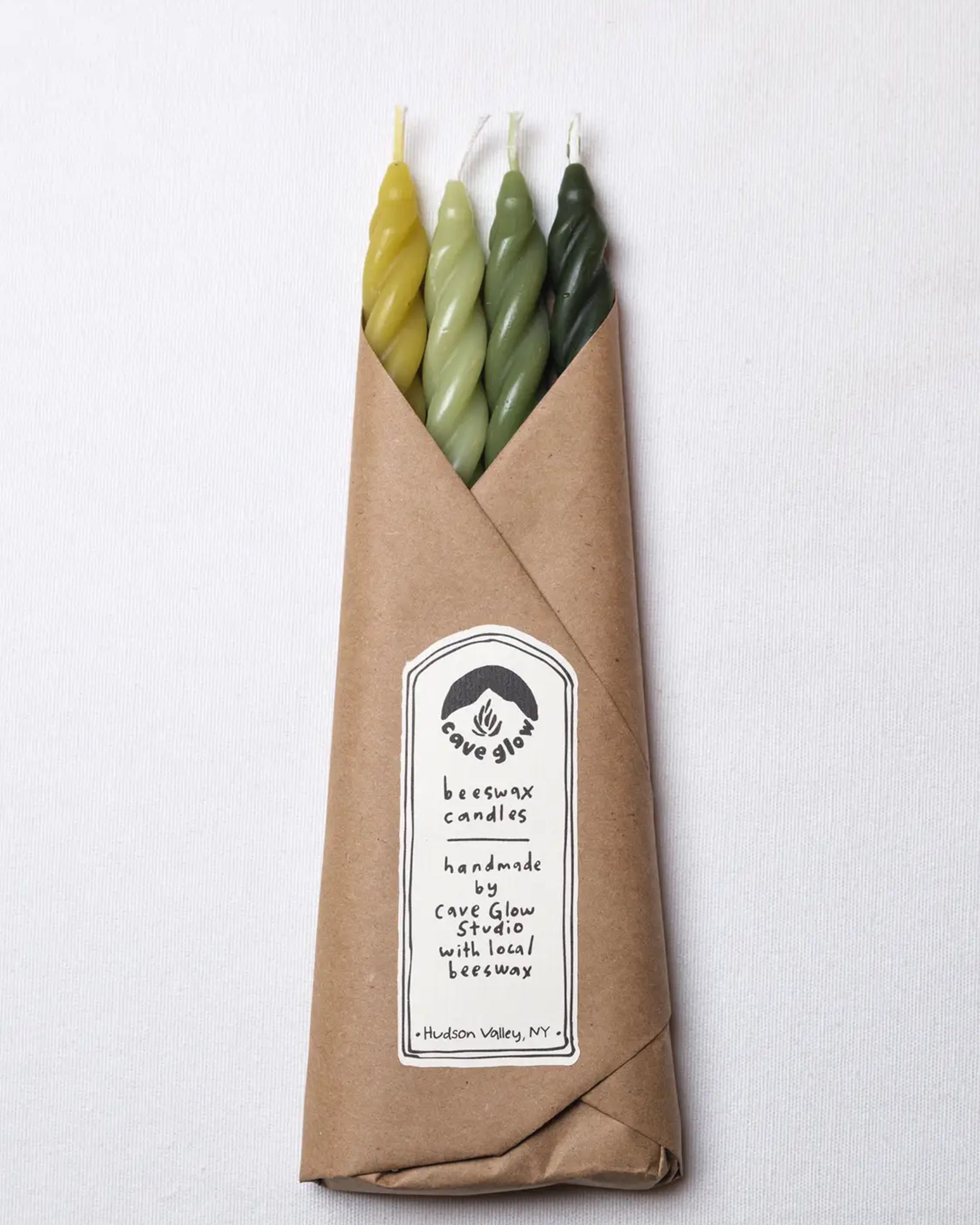 The Woodland Spiral Beeswax Candles S/4 by Faire are hand-crafted in yellow and green with natural dye, wrapped in brown paper, and labeled “handmade with local beeswax, Hudson Valley, NY.”.