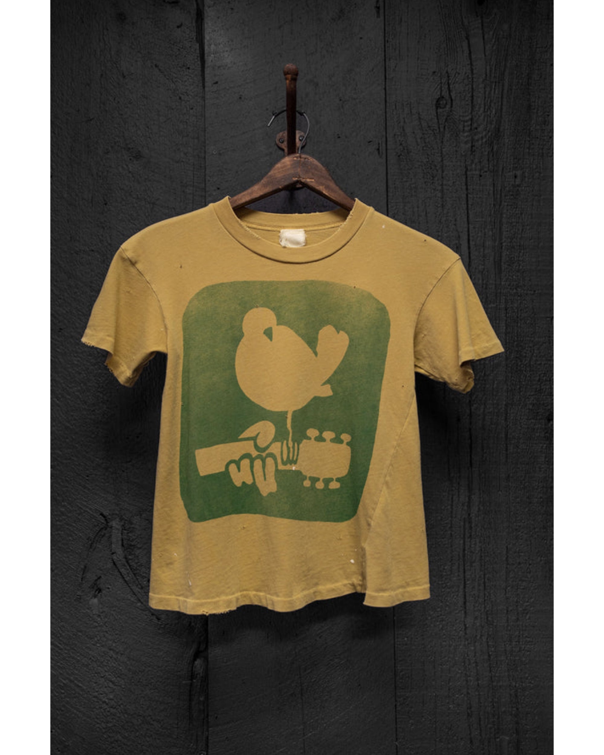 The Made Worn Woodstock Crop Tee Mustard is a vintage-fit, faded yellow t-shirt with a green bird graphic, shown on a wooden hanger against a dark textured wood background.
